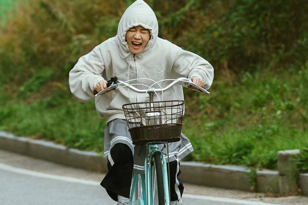 Seorang perempuan mengenakan hoodie abu-abu tertawa lepas saat mengayuh sepeda biru muda di jalan dengan latar pepohonan hijau.