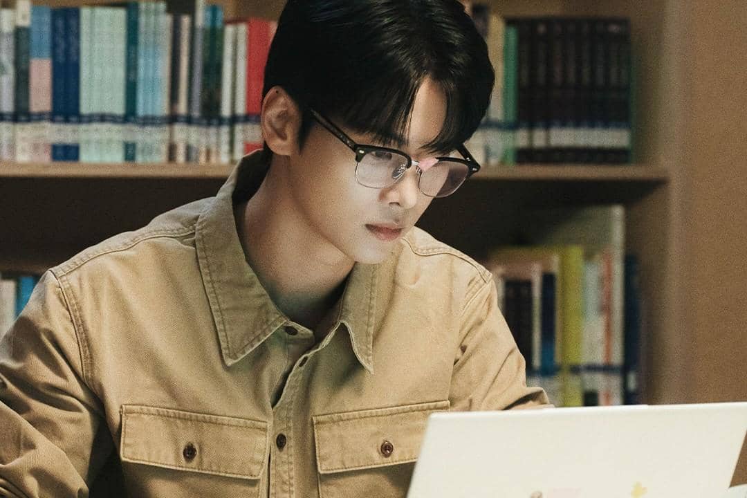 Seorang pria berkacamata mengenakan jaket krem sedang fokus menatap layar laptop di perpustakaan dengan rak buku di belakangnya.