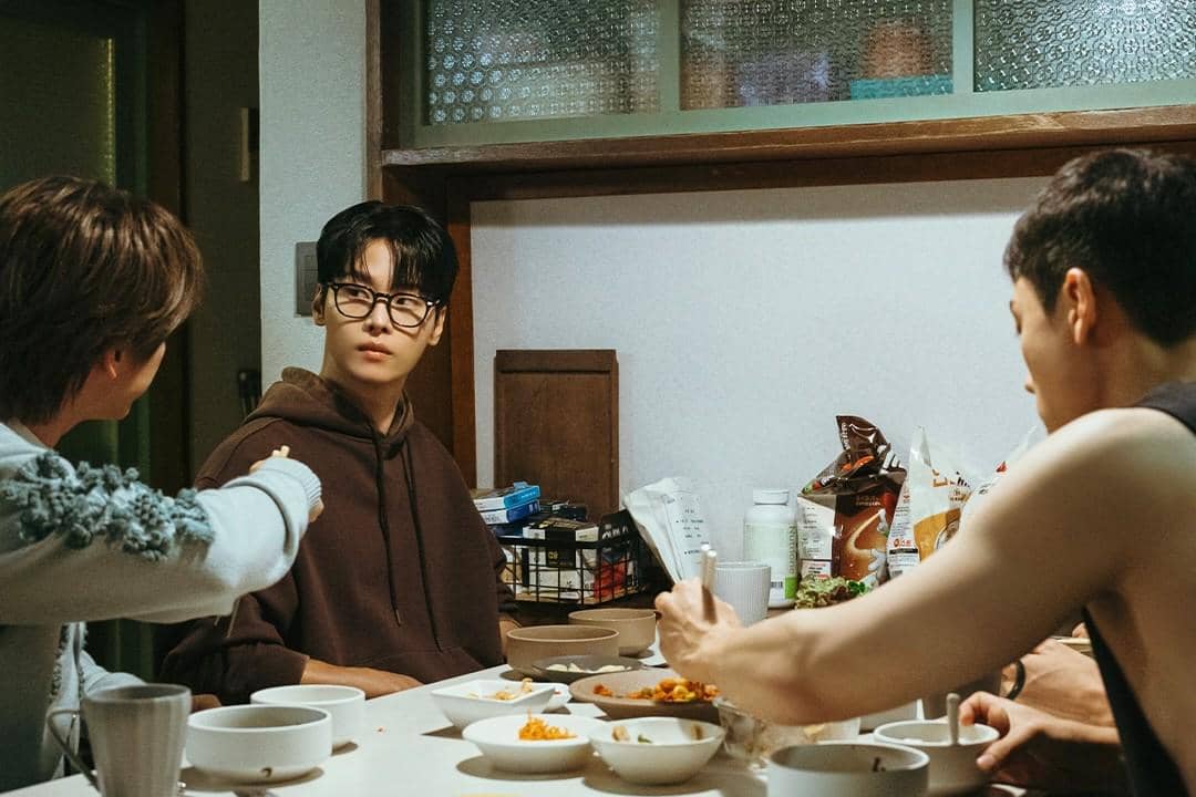 Tiga pria duduk di meja makan kecil dengan mangkuk dan makanan, salah satunya mengenakan hoodie cokelat dan kacamata.