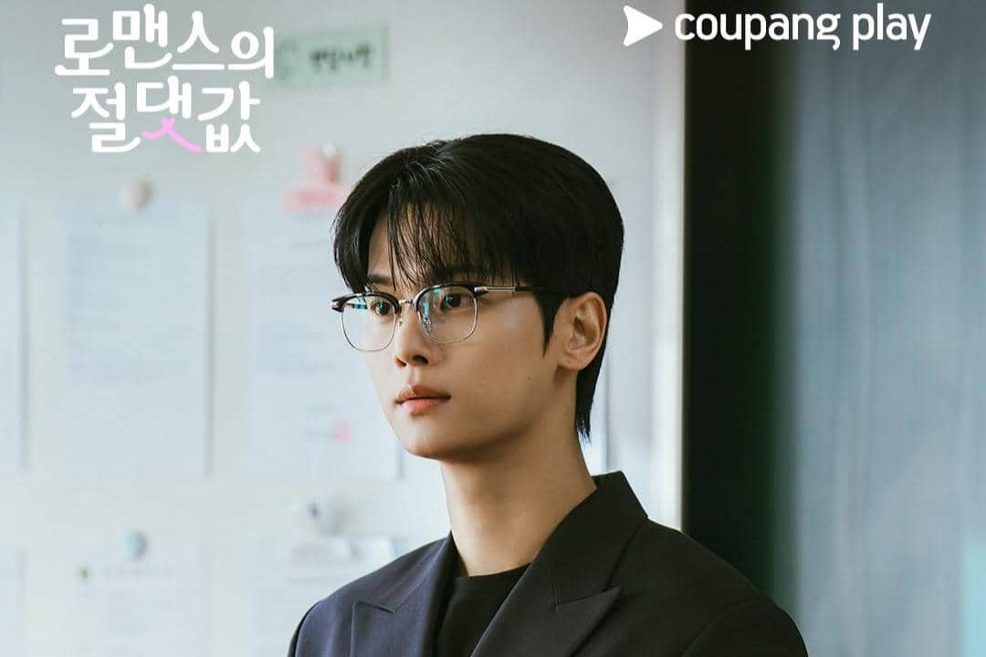 Seorang pria berpenampilan rapi mengenakan kacamata dan jas hitam berdiri di dalam ruangan dengan latar dinding berisi kertas tempel.