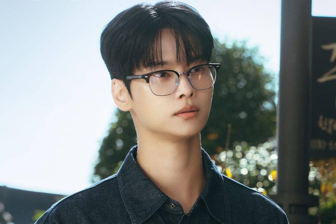 Seorang pria berambut hitam mengenakan kacamata dan kemeja denim berdiri di luar ruangan dengan latar pepohonan dan cahaya matahari.