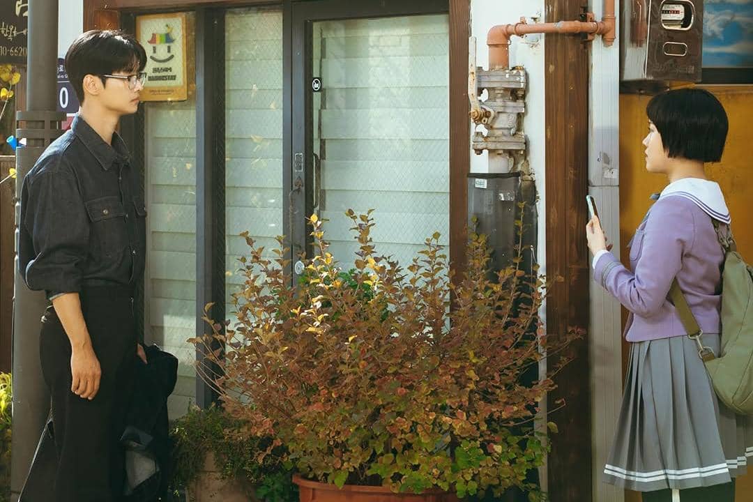 Seorang pria berjaket denim berdiri berhadapan dengan seorang gadis berseragam sekolah di depan pintu rumah dengan tanaman di depannya.