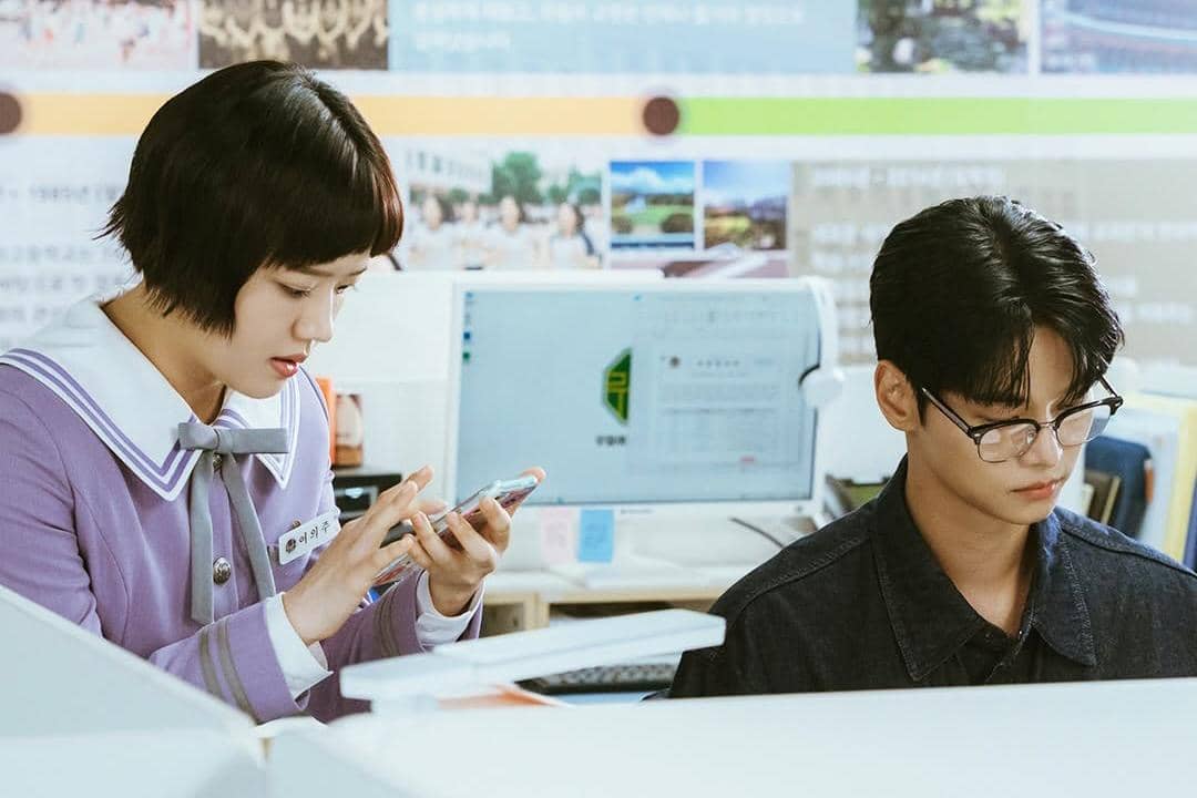 Dua orang sedang berada di kantor, seorang perempuan berseragam ungu melihat ponsel sementara pria berkacamata fokus di depan komputer.