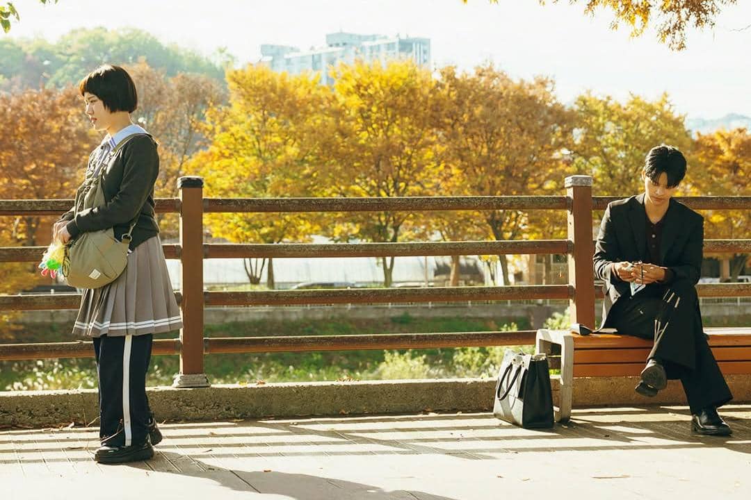 Seorang perempuan berseragam sekolah berdiri di tepi sungai sementara seorang pria duduk di bangku taman dengan latar pepohonan musim gugur.