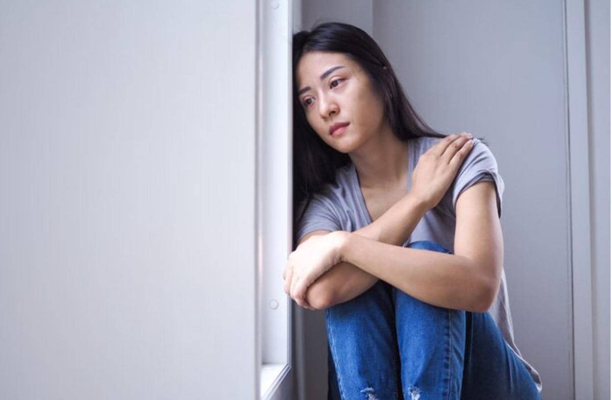 Perempuan duduk bersandar di dekat jendela dengan ekspresi sedih dan menyesal, mengenakan kaus abu-abu dan celana jeans biru.