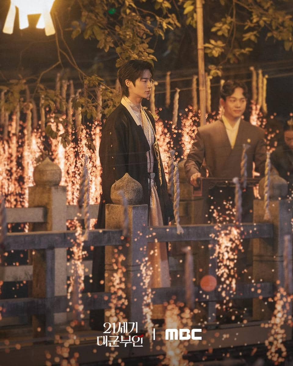 Dua aktor berdiri di jembatan kayu dengan percikan api di sekelilingnya dalam adegan drama Perfect Crown yang bernuansa malam.