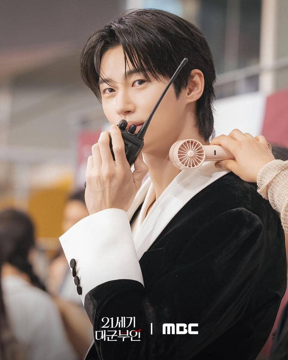 Seorang aktor mengenakan jas hitam dan putih memegang walkie-talkie sambil diarahkan kipas kecil di lehernya di lokasi syuting drama Perfect Crown.