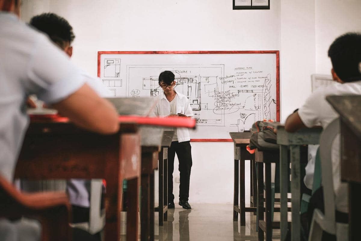 Seorang siswa berdiri di depan kelas menjelaskan materi di papan tulis berisi gambar teknik, sementara teman-temannya duduk memperhatikan.