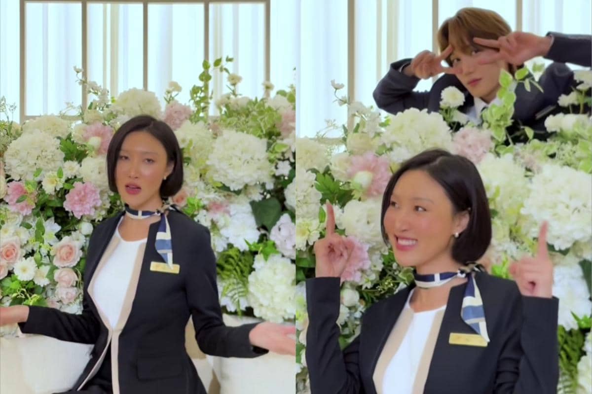 Seorang wanita berambut pendek mengenakan setelan hitam dan syal bermotif berpose di depan rangkaian bunga putih dan merah muda, dengan seorang pria berpose lucu di belakangnya.