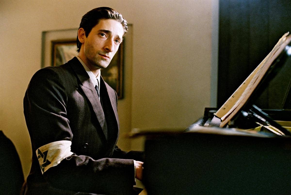 Seorang pria mengenakan setelan jas gelap duduk di depan piano dengan lembaran musik di atasnya dalam adegan film The Pianist.