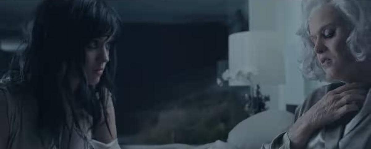 Dua perempuan duduk di ruangan redup, satu berambut hitam dan satu berambut putih, tampak berbicara dengan ekspresi emosional.