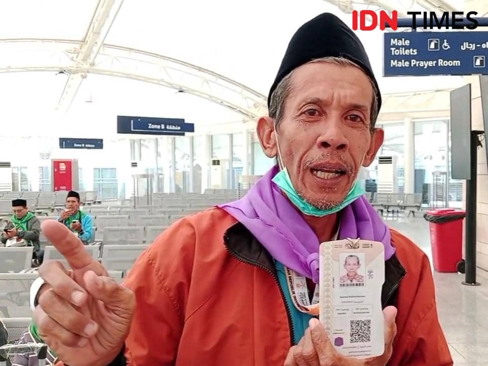 Seorang jemaah haji mengenakan jaket oranye dan syal ungu menunjukkan kartu identitas di area kedatangan bandara Madinah, Arab Saudi.