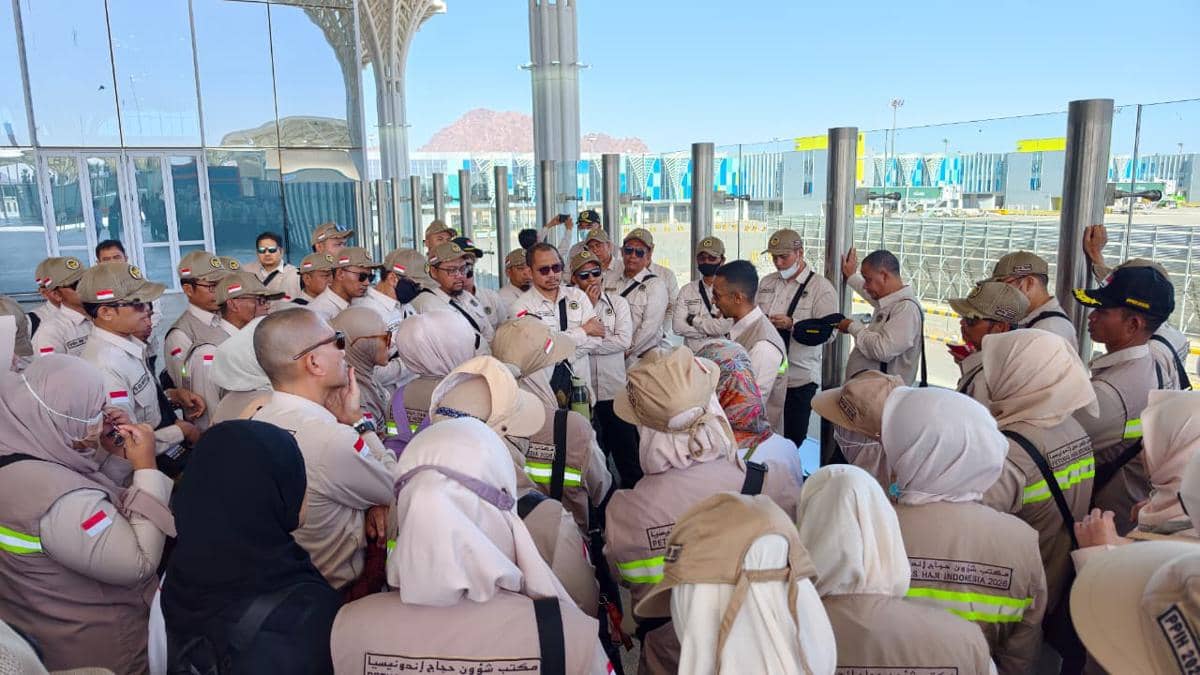 Petugas haji Indonesia mengenakan seragam krem berkumpul di area luar bandara dengan latar bangunan modern dan pegunungan.