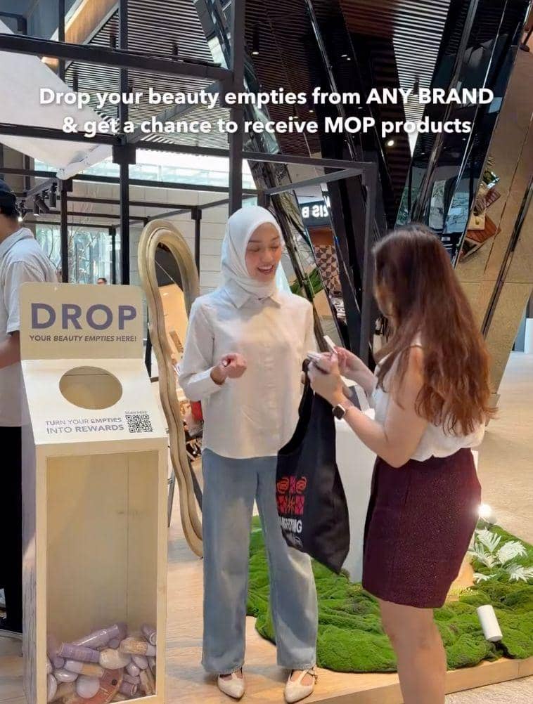Dua wanita berinteraksi di area publik dengan kotak daur ulang bertuliskan 'Drop your beauty empties here' untuk program keberlanjutan MOP Beauty.