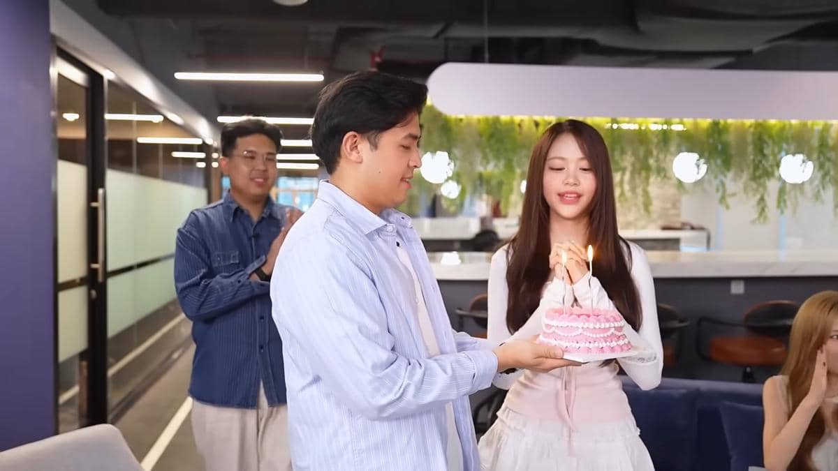 Jerome Polin memberikan kue ulang tahun berwarna merah muda dengan lilin menyala kepada Carmen di ruang kantor modern.