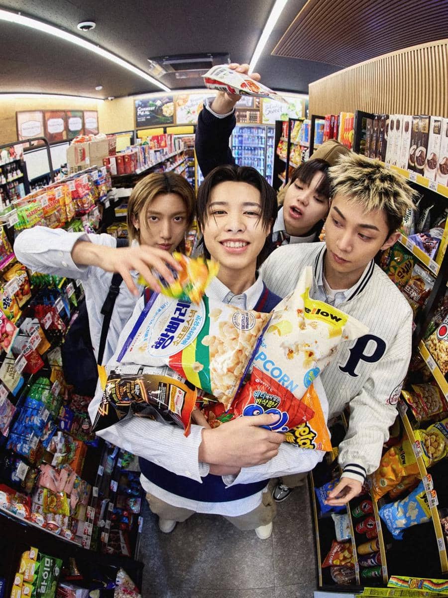 Empat anggota grup musik LNGSHOT tersenyum sambil memegang berbagai camilan di dalam sebuah minimarket penuh warna.