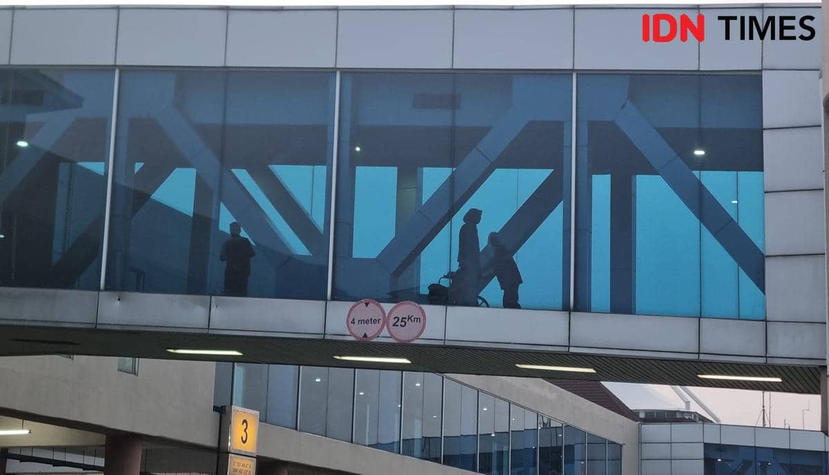 Jemaah prioritas berjalan melalui garbarata di Bandara Sultan Mahmud Badaruddin II Palembang dengan latar kaca biru dan struktur baja.