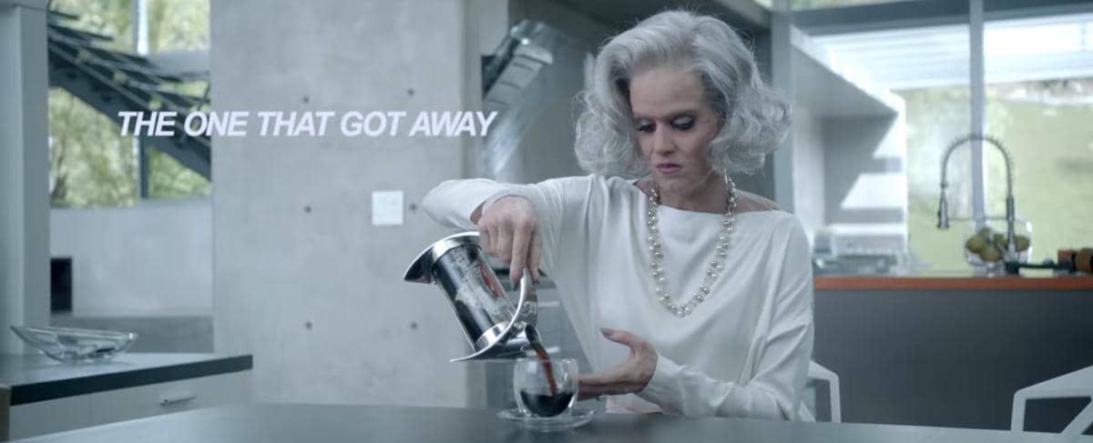 Seorang wanita berambut abu-abu mengenakan blus putih menuangkan kopi ke dalam cangkir di dapur modern dengan pencahayaan alami.