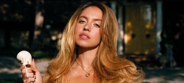 Sydney Sweeney memegang es krim di luar ruangan dengan latar rumah berpintu kuning di belakangnya.
