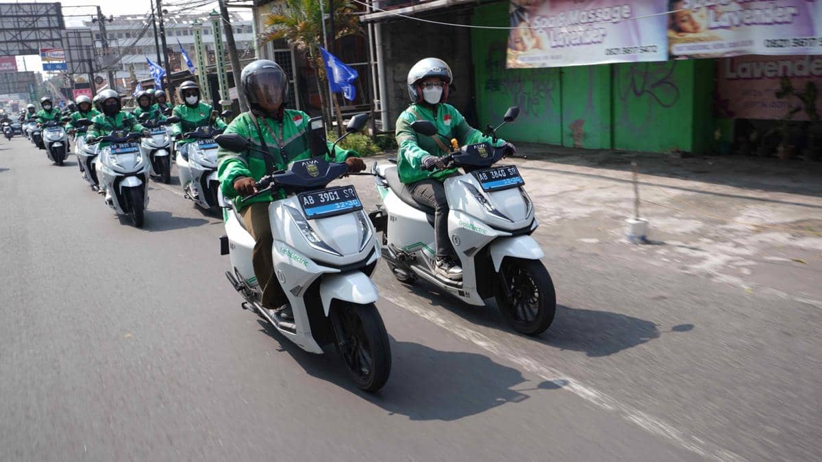 Sejumlah pengendara ojek online mengenakan jaket hijau mengendarai motor listrik berwarna putih melaju di jalan perkotaan.