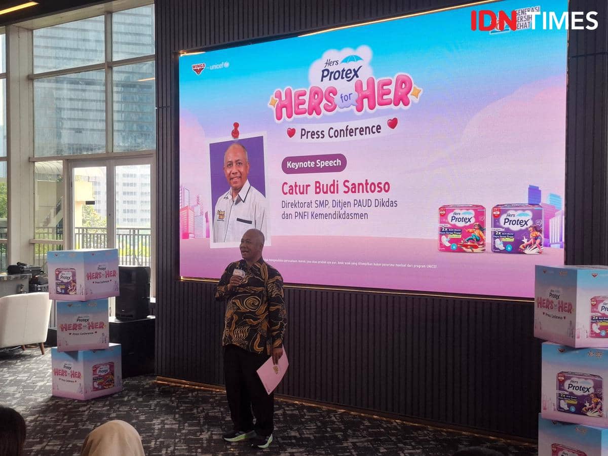 Seorang pembicara memegang mikrofon di depan layar besar bertema Hers Protex for Her dalam konferensi pers di Jakarta Selatan.