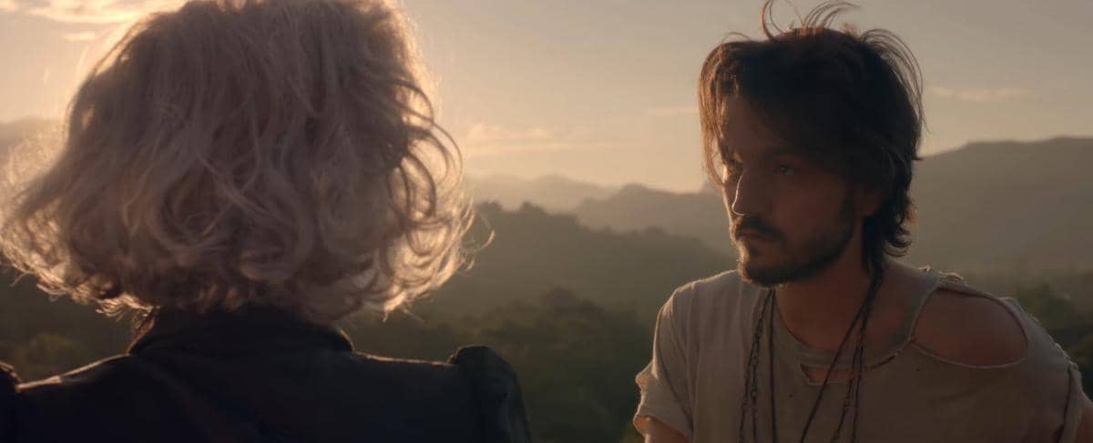 Seorang pria berambut panjang menatap wanita berambut pirang pendek di luar ruangan saat matahari terbenam dengan latar pegunungan.