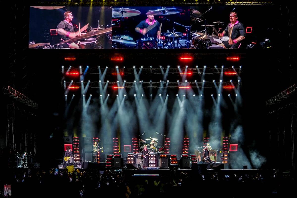 Panggung konser Green Day di Jakarta dengan pencahayaan warna-warni, layar besar menampilkan drummer, dan penonton memenuhi area depan.