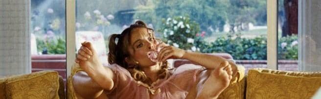 Sydney Sweeney mengenakan gaun berwarna merah muda duduk santai di sofa sambil memegang permen di mulutnya di ruang tamu yang terang.