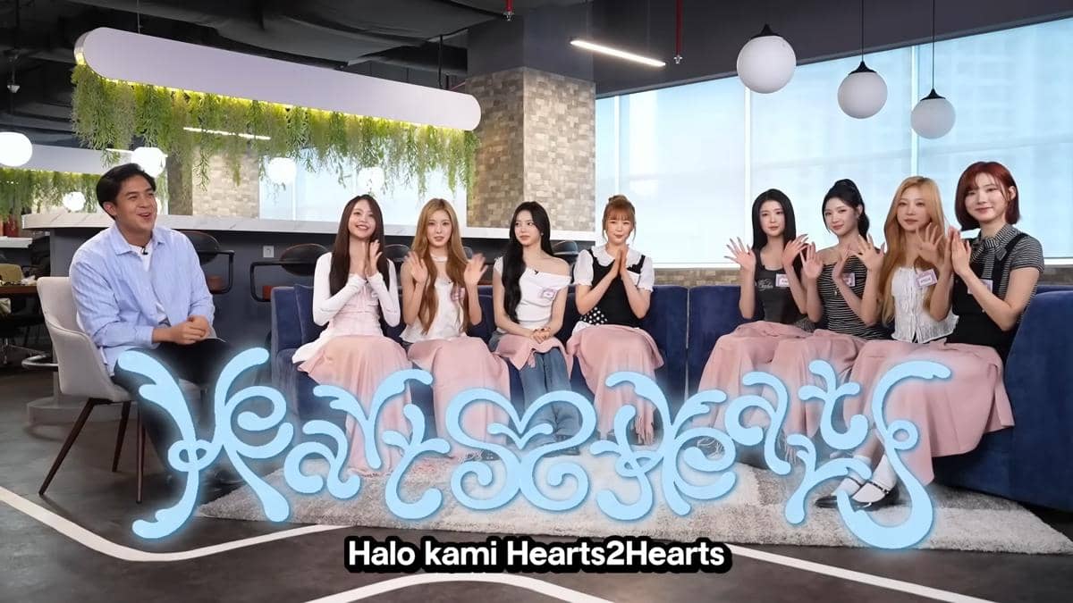 Jerome Polin duduk bersama delapan anggota grup Hearts2Hearts di ruang modern dengan dekorasi hijau gantung dan logo besar di depan mereka.