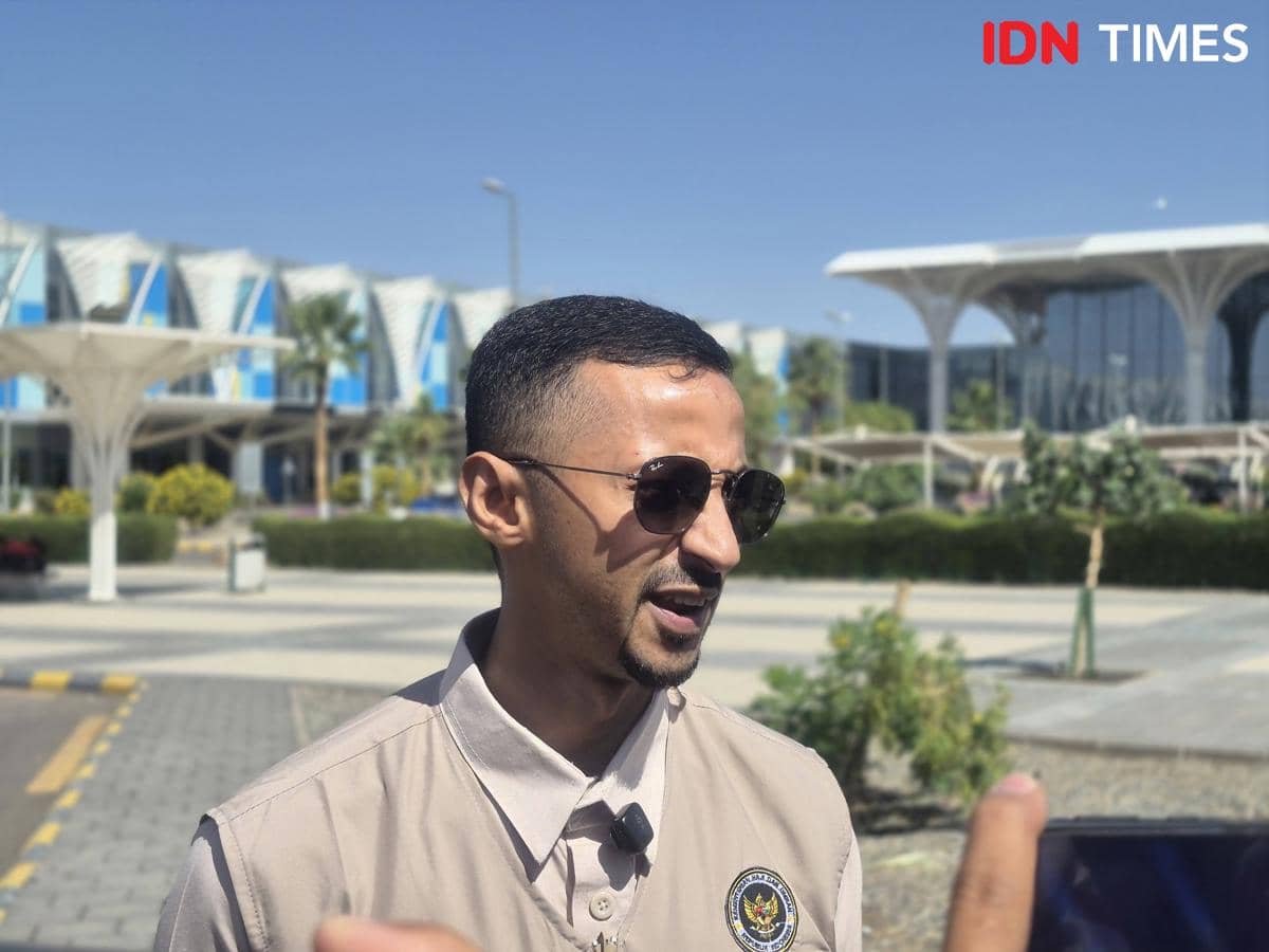 Seorang pria mengenakan kemeja krem dan kacamata hitam berbicara di area luar ruangan dengan latar gedung modern dan langit cerah.