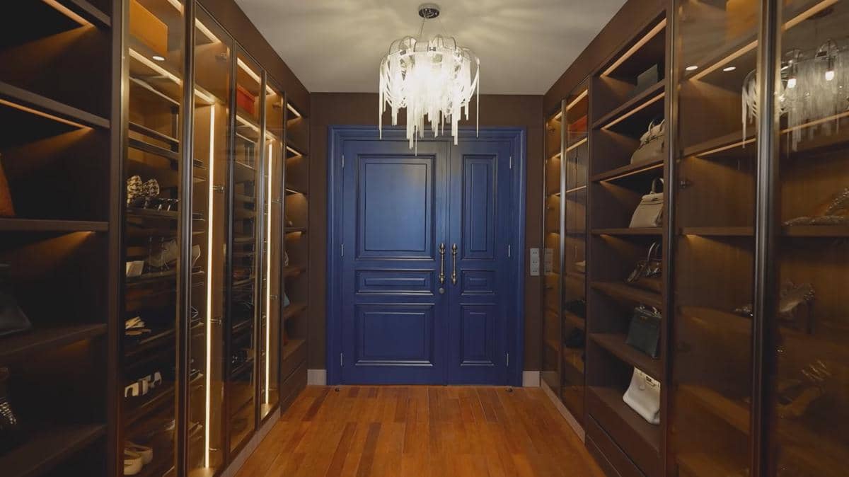 Ruang lemari pakaian mewah dengan rak kayu berlampu, pintu ganda berwarna biru tua, dan lampu gantung kristal di langit-langit.