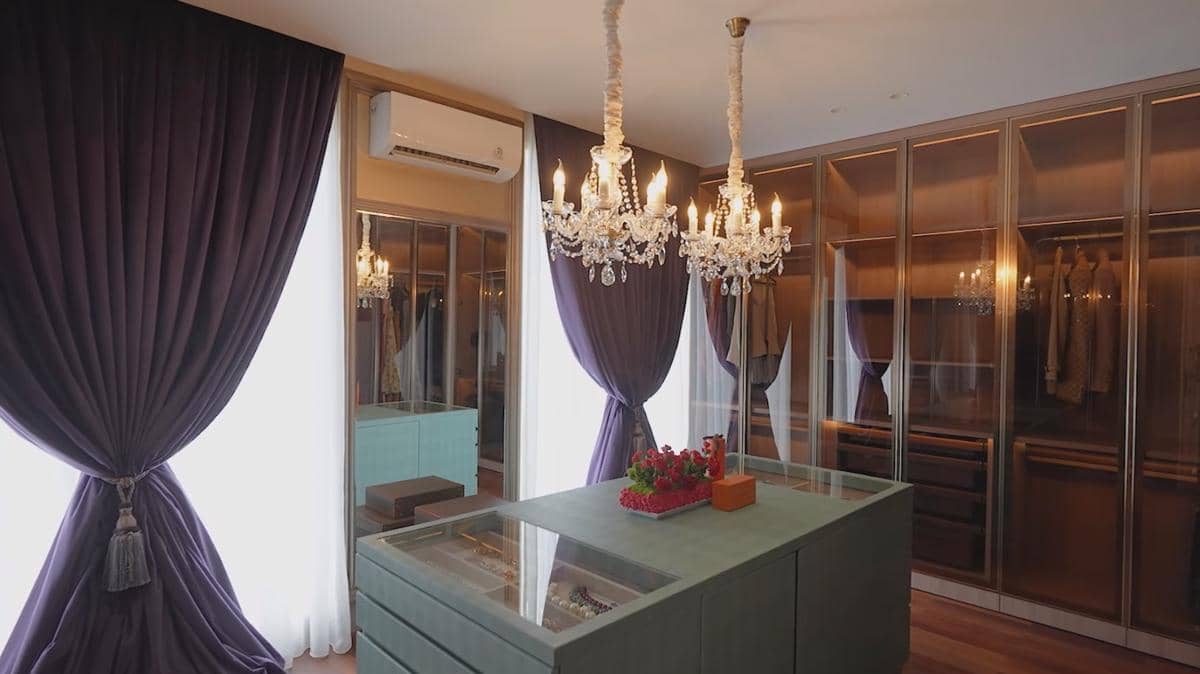 Ruang ganti mewah dengan lemari kaca besar, tirai ungu tebal, lampu gantung kristal, dan meja pajangan berlapis kaca di tengah ruangan.