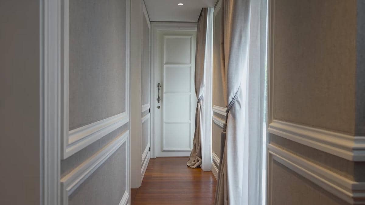 Lorong kamar bergaya mewah dengan dinding berpanel putih, tirai abu-abu lembut, dan lantai kayu yang elegan menuju pintu putih.