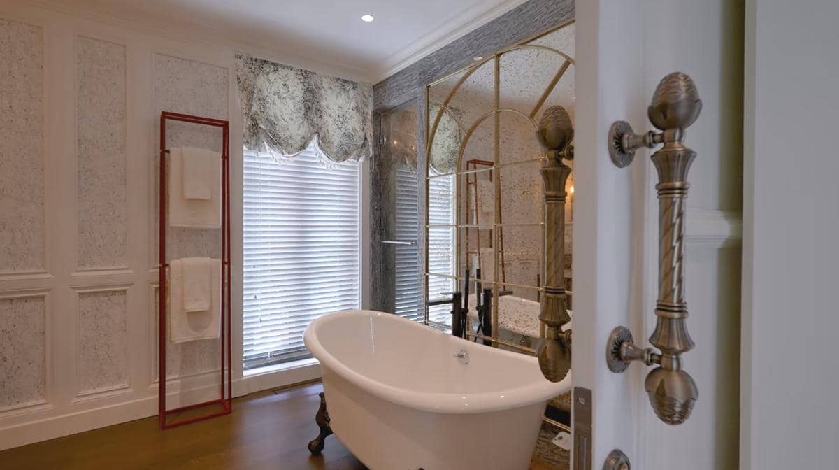 Kamar mandi mewah dengan bathtub berdiri, cermin besar berbingkai emas, dan jendela dengan tirai bermotif elegan.