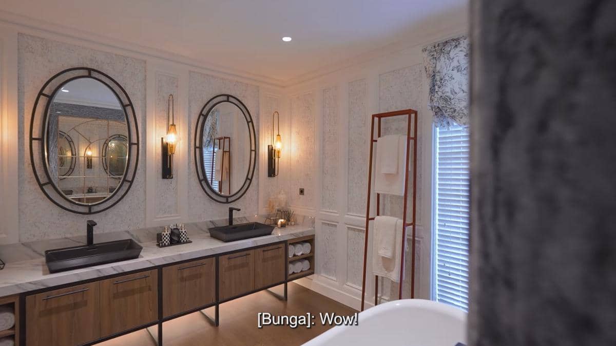 Kamar mandi mewah dengan dua wastafel hitam, cermin oval besar, pencahayaan hangat, dan rak handuk di dekat jendela ber tirai.