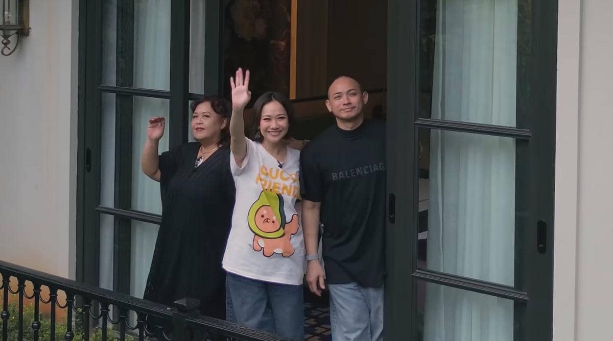 Tiga orang berdiri di balkon kamar luas dengan pintu kaca besar, salah satunya melambaikan tangan sambil tersenyum ke arah luar.