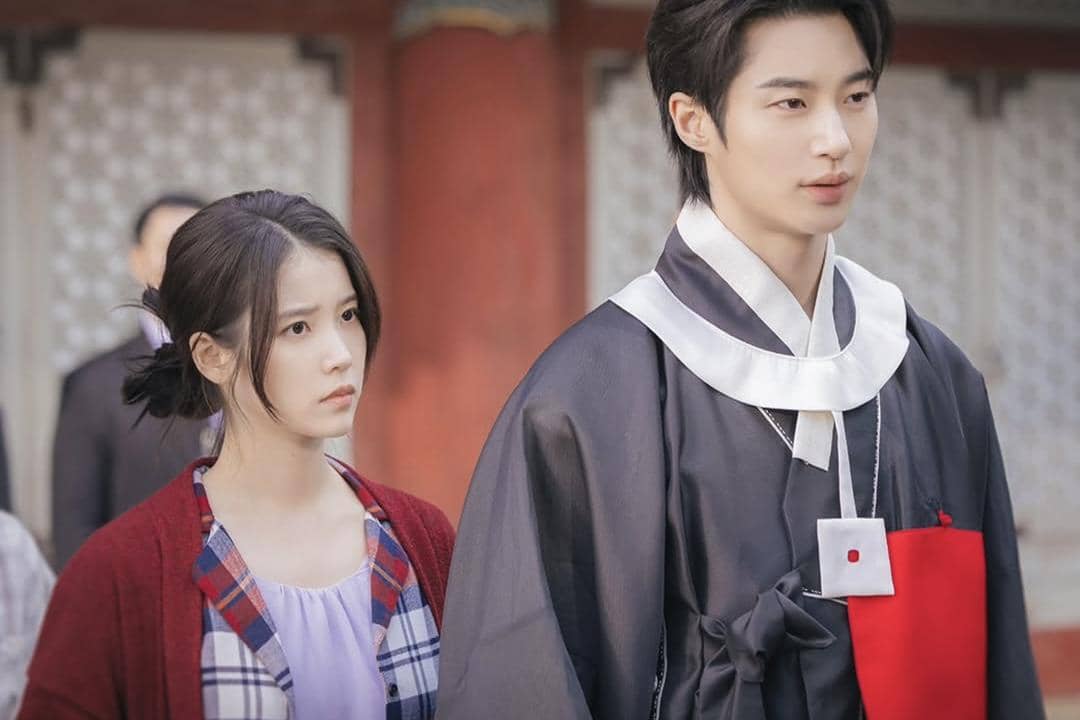 Dua karakter dalam drama Korea Perfect Crown berdiri berdampingan, satu mengenakan hanbok hitam dan satu berpakaian kasual dengan ekspresi serius.