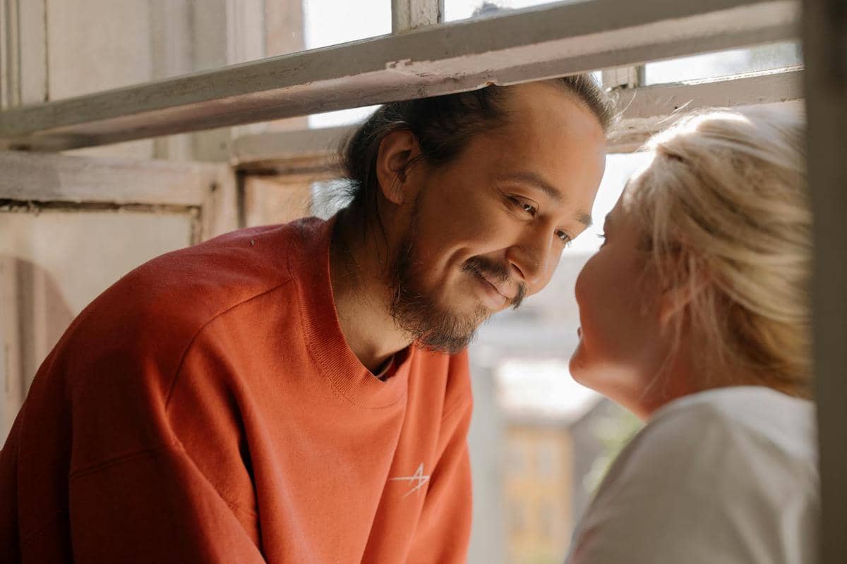 Seorang pria tersenyum sambil menatap wanita di dekat jendela terbuka dengan suasana hangat dan akrab di siang hari.