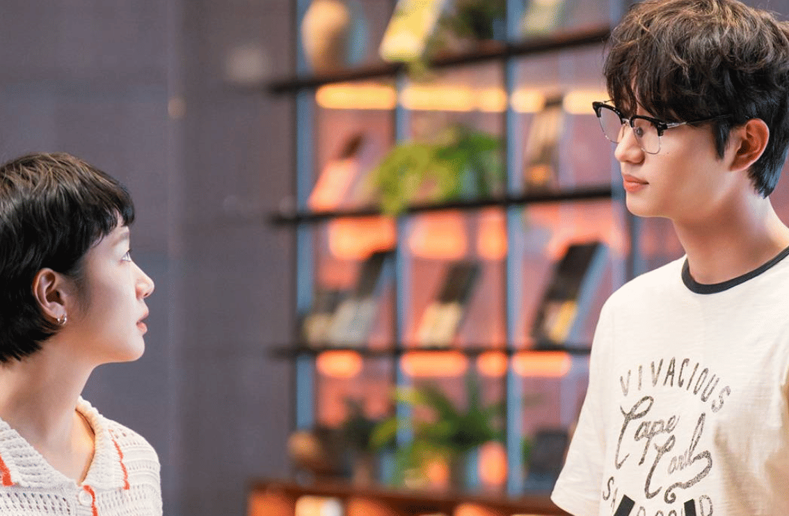 Dua karakter dalam drama Korea Yumi's Cells 3 berdiri saling berhadapan di ruangan dengan latar rak buku dan pencahayaan hangat.