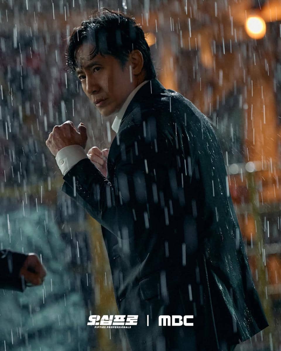 Seorang pria mengenakan setelan jas basah berdiri dalam posisi bertarung di bawah hujan deras pada adegan drama Fifties Professionals.