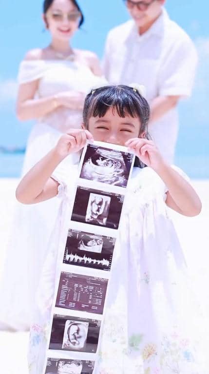 Nadlyne tersenyum sambil memegang hasil USG di pantai dengan dua orang dewasa berdiri di latar belakang mengenakan pakaian putih.