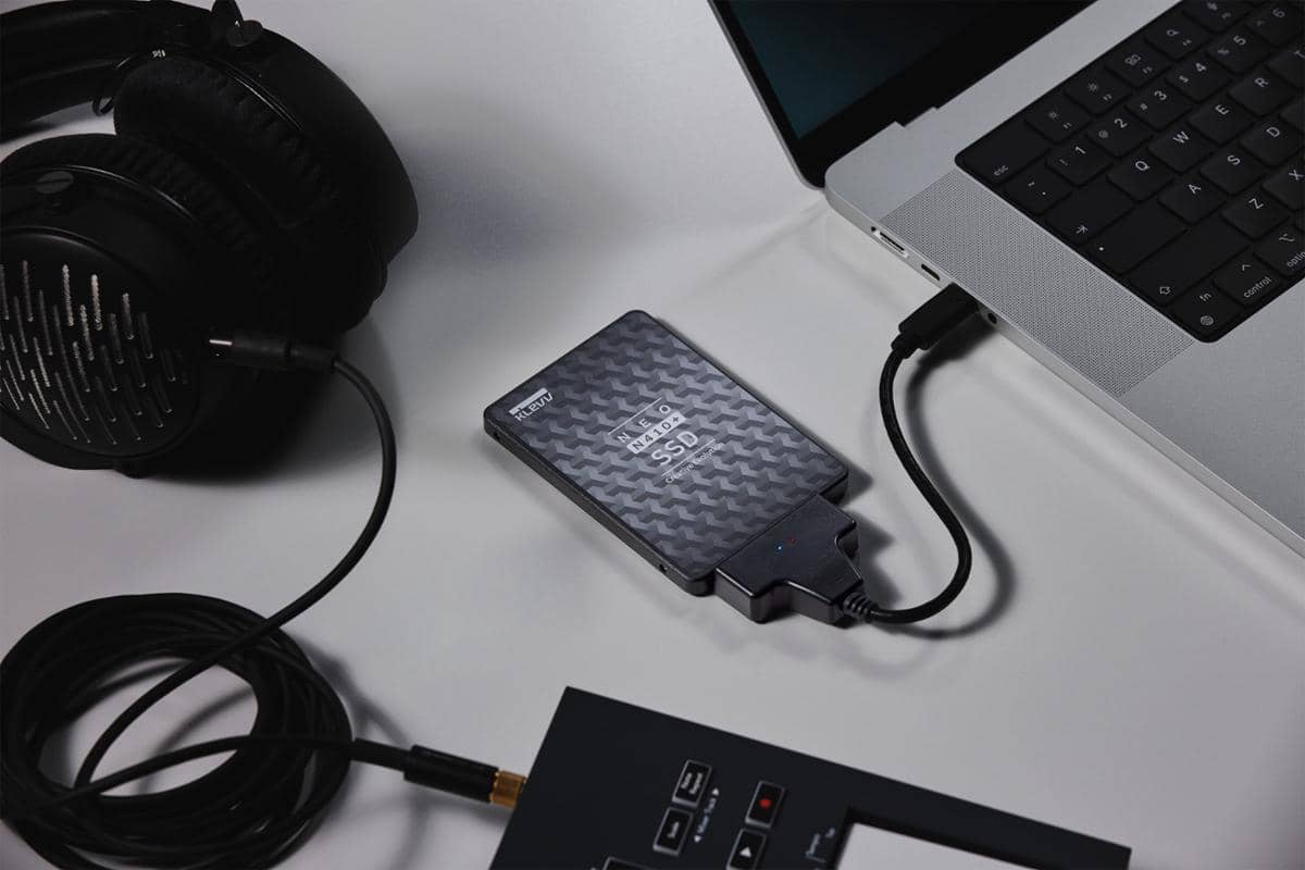 SSD Klevv Neo N410+ terhubung ke laptop melalui kabel USB di meja bersama headphone dan perangkat audio lainnya.