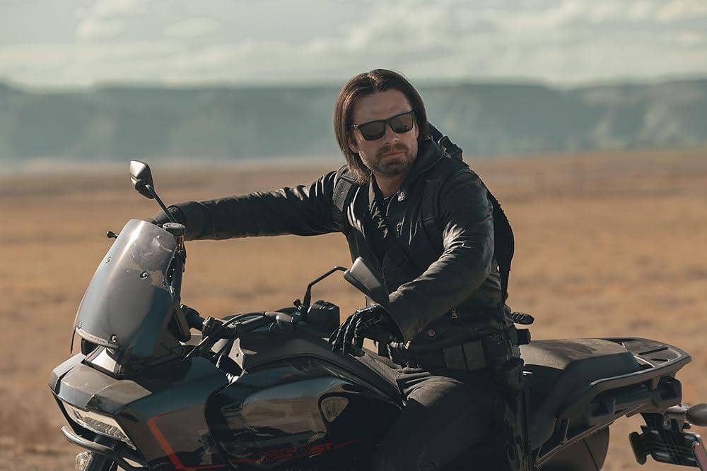 Bucky barnes berjaket hitam dan berkacamata hitam mengendarai motor di area padang luas dengan latar pegunungan di kejauhan.