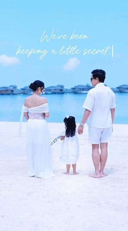 Keluarga Nanda Arsyinta berdiri di pantai bersama suami dan anak kecil mereka, mengenakan pakaian putih dengan latar laut biru jernih.