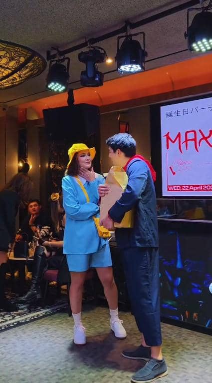 Luna Maya mengenakan kostum biru dengan topi kuning memberi hadiah ke Maxime di acara ulang tahun di dalam ruangan.