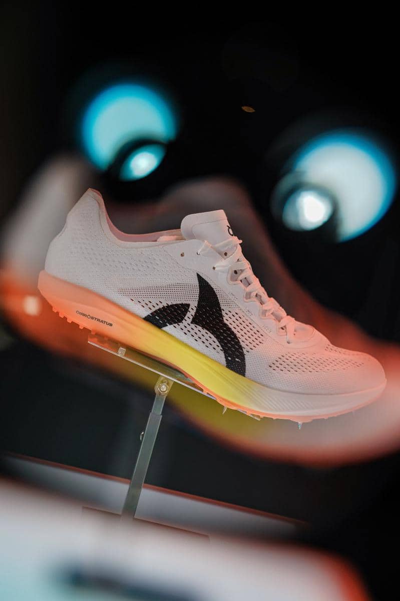 Sepatu olahraga Ortuseight berwarna putih dengan sol gradasi kuning-oranye dipamerkan di atas dudukan logam dengan latar gelap.