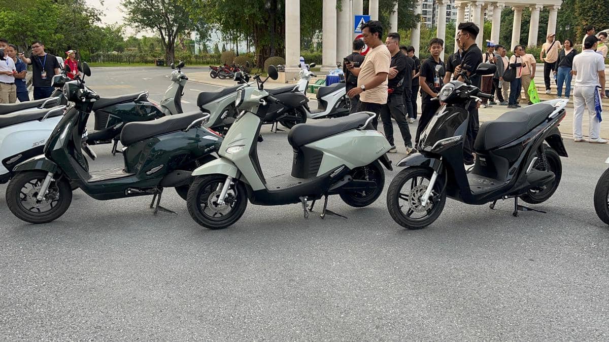 Beberapa skuter Vinfast berwarna hitam, hijau, dan abu-abu diparkir berjajar di area terbuka dengan sekelompok orang di latar belakang.