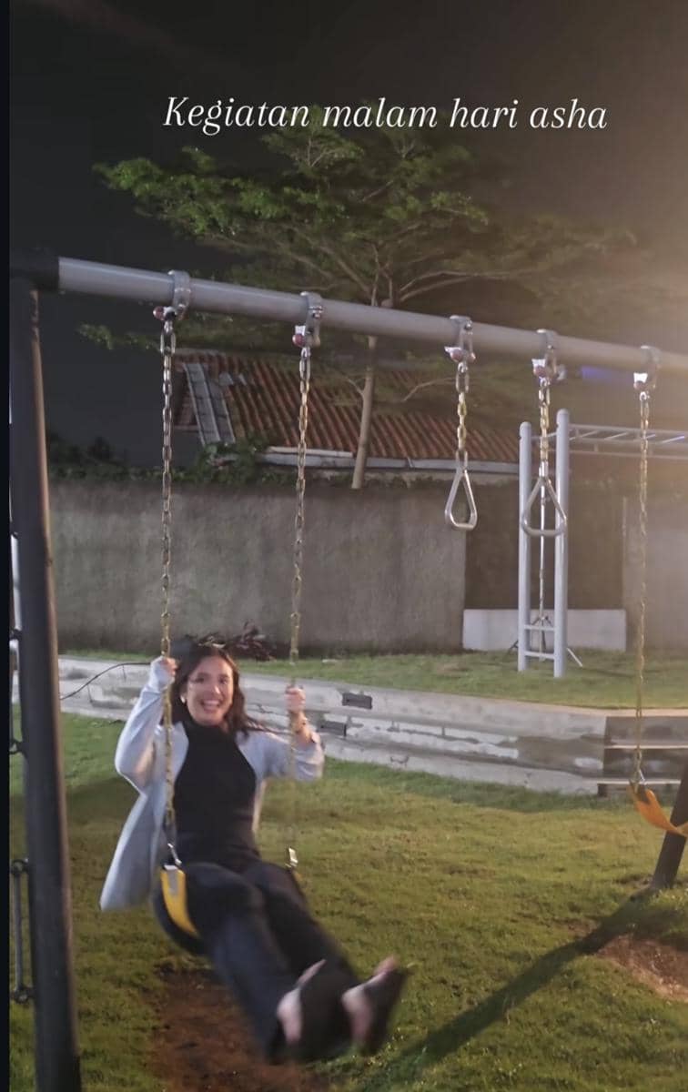 Seorang wanita tersenyum sambil bermain ayunan di malam hari di area rumput dengan latar belakang tembok dan pepohonan.