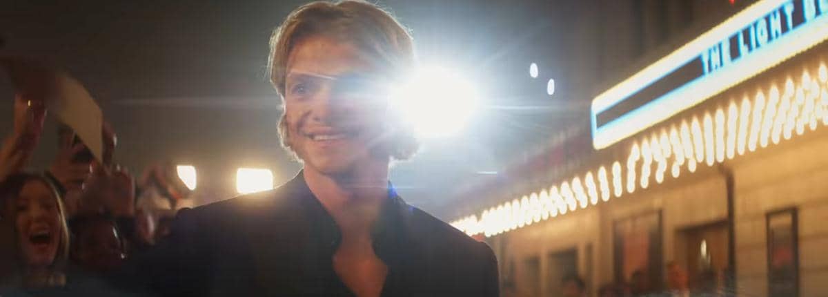 Matt Hagen tersenyum di depan gedung teater dengan lampu terang dan kerumunan penggemar di latar belakang pada malam hari.