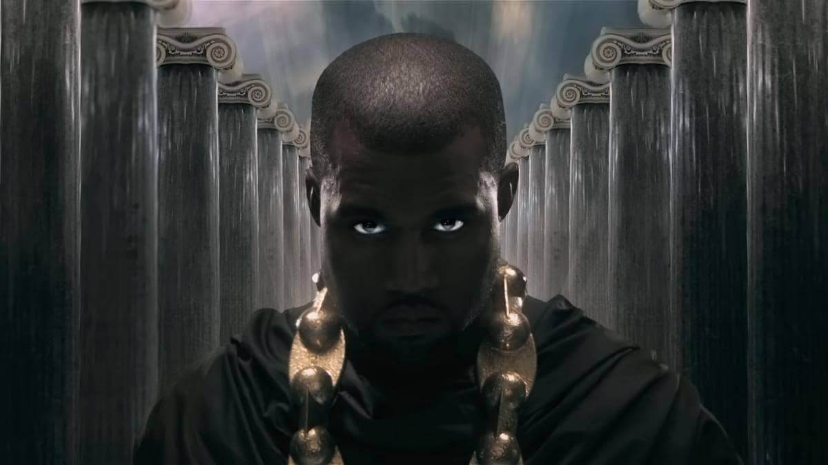 Kanye West mengenakan jubah hitam dan kalung besar berwarna emas, berdiri di antara deretan pilar bergaya klasik.